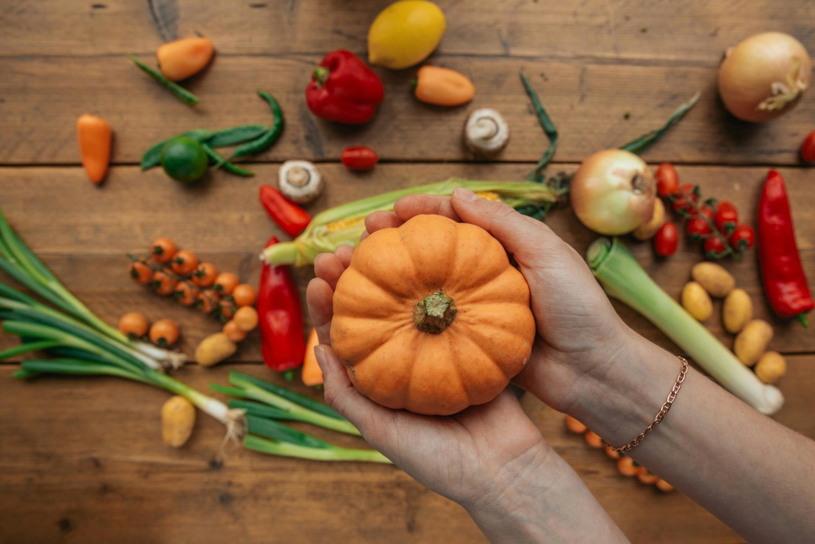 Hands holding a mini pumpkin with assorted fresh vegetables scattered on a wooden table.